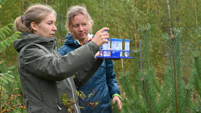 Two women take geological samples outdoors. In the background are young trees.