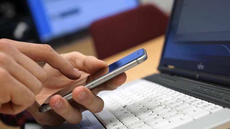 Person sitzt vor dem Notebook und tippt auf seinem Handy