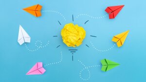 Bunte Papierflieger fliegen aus verschiedenen Richtungen auf eine gelbe Papierkugel in der Mitte zu, die symbolisch als Glühbirne dargestellt ist – auf blauem Hintergrund. Die Szene steht für kreative Ideen und gemeinsames Denken.