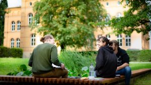 Drei junge Studierende sitzen in einem Garten auf einer Bank und unterhalten sich
