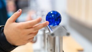 Eine Hand berüht eine blaue Kugel auf einer Schiene und bringt sie zum Rollen
