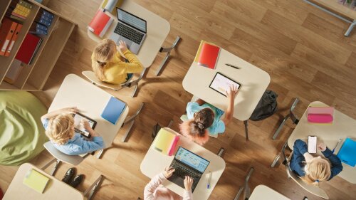 Mehrere Kindern sitzen an Einzeltischen in einem Schulzimmer und arbeiten an Laptops, Tablets oder Smartphones.