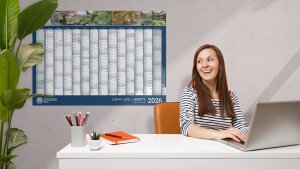 Eine Frau sitzt lächelnd am Schreibtisch. Hinter ihr hängt der Wandkalender 2026 der Uni Jena.