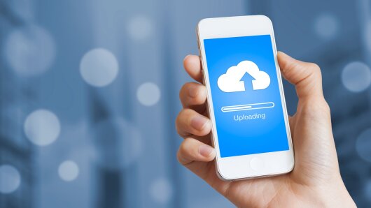 Eine Hand hält ein Smartphone, auf dem das Wort »Uploading« steht.