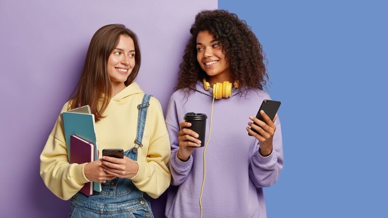 Zwei junge Frauen tragen pastellfarbene Hoodies und lächeln sich an.