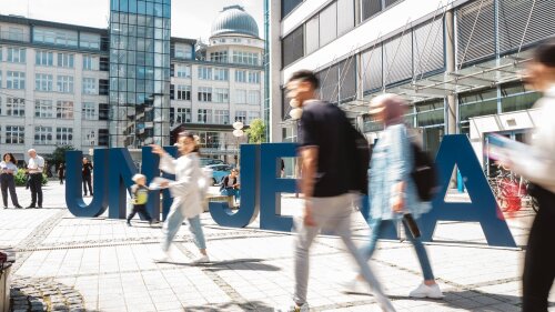 Vor den großen Uni-Jena-Buchstaben auf dem Ernst-Abbe Platz sind unscharf Personen zu erkennen.