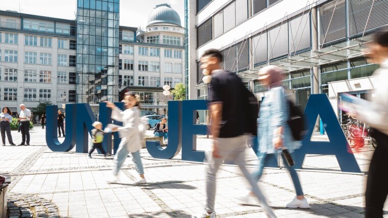Vor den großen Uni-Jena-Buchstaben auf dem Ernst-Abbe Platz sind unscharf Personen zu erkennen.