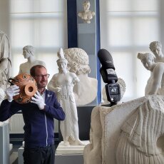 Dr. Dennis Graen hält, umringt von Gips-Skulpturen, eine antike Schale und lächelt in die Kamera.