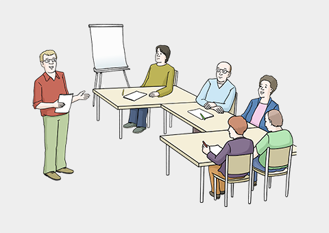 Eine Gruppe von Männern und Frauen sitzen an einem langen Tisch. Vor ihnen steht ein Mann mit einem Zettel in der Hand. Neben dem Tisch steht auch ein Flipchart. Das ist eine Tafel mit einem großem Schreibblock.