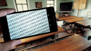 Text in Binärcode auf dem Display eines Smartphones, im Hintergrund das Klassenzimmer einer historischen Dorfschule