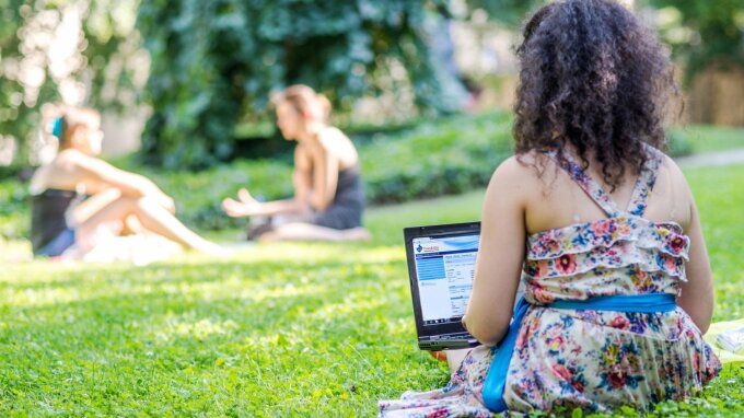 Student sitting on a meadow with her back to the camera and working on the laptop