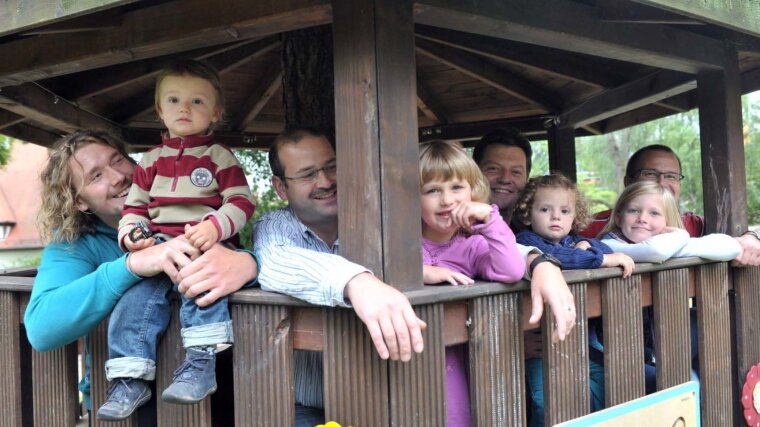 Vier Kinder und ihre Väter lachen aus einem Kinderhaus auf einem Spielplatz.