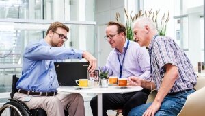 Mitarbeiter im Rollstuhl sitzt mit zwei Kollegen in der Cafeteria und zeigt eine Präsentation auf seinem Notebook-Monitor