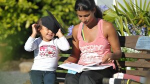 Mother and toddler on a bench in a park—the mother is reading, the toddler is playing with a mortarboard